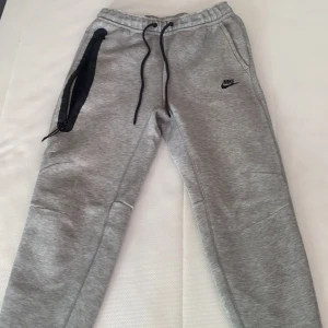 Grå mjukisbyxor från Nike - Snygga grå mjukisbyxor från Nike med svart logga på sidan. Byxorna har en elastisk midja med snörning och praktiska fickor. Perfekta för en avslappnad stil. lite noppriga på baksidan