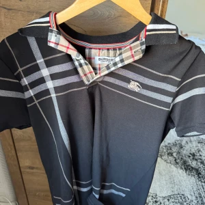 Svart t-shirt från Burberry - Snygg svart t-shirt från Burberry med klassiskt rutigt mönster på kragen. Den har en diskret logga på bröstet och är kortärmad. Perfekt för en stilren look.