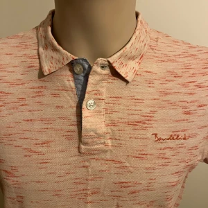 Rosa pikétröja från Bondelid - Säljer en snygg rosamelerad pikétröja från Bondelid med röda detaljer. Tröjan har en klassisk krage och knappar framtill. Perfekt för en avslappnad stil.