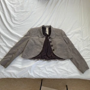 Grå och brun rutig kavaj/kofta  - Säljer en elegant och vintage grå och brun kavaj/kofta med rutigt mönster och bruna knappar framtill. Kavajen har en klassisk krage och är kortare i modellen, samt har en puffig baksida för en mer feminiserad look. Perfekt för att lyfta din outfit med stil✨