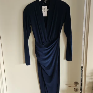 Mörkblå klänning med slit  - Elegant mörkblå klänning med draperad design och djup v-ringning. Klänningen har långa ärmar och en smickrande passform som passar perfekt för festliga tillfällen.