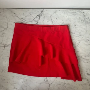 Röd kjol från Pull&Bear - Snygg röd kjol från Pull&Bear med volangdetaljer. Perfekt för att ge en färgklick till din outfit. Kjolen har en omlott-design som ger en trendig look.