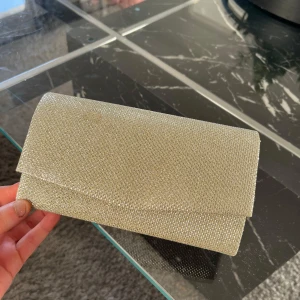 Glittrig kuvertväska från Boohoo - Säljer en snygg glittrig kuvertväska i guld från Boohoo. Perfekt för att ge din outfit lite extra bling. Väskan har en magnetknäppning. Den är kompakt men rymmer det viktigaste för en kväll ute. 👜✨obs kedjan är tyvärr borta! Har en liten fläck på väskan i vissa ljus