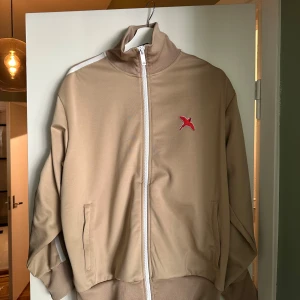 Beige zipper från Axel Arigato - Säljer en beige jacka från Axel Arigato med vit dragkedja och en röd broderad logga på bröstet. Jackan har långa ärmar med vita ränder längs sidorna och ribbade muddar vid ärmslut och nederkant. Perfekt för en stilren look!