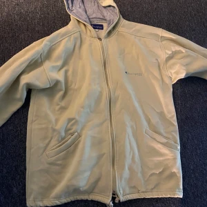 Beige/gul jacka/zip hoodie från Champion - Säljer en beige jacka från Champion med dragkedja och huva. Jackan har två fickor framtill och ett stort Champion-tryck på ryggen. Perfekt för en avslappnad stil. 