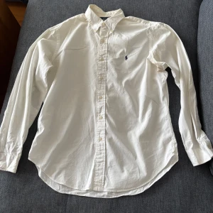 Vit skjorta från Ralph Lauren - Klassisk vit skjorta från Ralph Lauren i bomull med broderad logga på bröstet. Skjortan har en normal passform och långa ärmar med knappar. Perfekt för en stilren look.