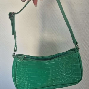Grön axelväska i syntet - Snygg grön axelväska med krokodilpräglat mönster. Väskan har en justerbar axelrem och en praktisk dragkedja upptill. Perfekt för att ge en färgklick till din outfit!