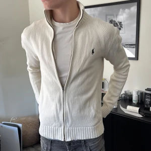 Vit kofta från Polo Ralph Lauren - Säljer en stilren vit kofta från Polo Ralph Lauren med dragkedja och ribbade detaljer. Perfekt för en avslappnad och snygg look. Den har långa ärmar och en liten logga på bröstet. Passar bra till jeans eller chinos.