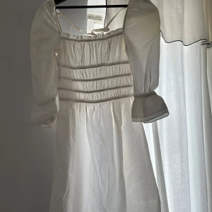 Vit klänning med puffärmar - Charmig vit klänning med puffiga trekvartsärmar och rynkade detaljer på överdelen. Perfekt för en somrig look med sin luftiga design och romantiska stil. Klänningen har en smickrande passform och är tillverkad i ett lätt material.