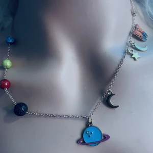 Made by prisförslag- Färgglatt halsband med planetmotiv - Unikt halsband med färgglada pärlor som representerar planeter och hängsmycken i form av en måne och stjärna. Halsbandet har en kedja i metall och ett centralt blått planetmotiv med ringar. Perfekt för den som älskar rymden och vill ha ett lekfullt smycke.