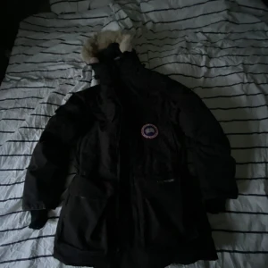 Svart parkajacka från Canada Goose - Säljer en svart parkajacka från Canada Goose med pälsdetaljer på huvan. Jackan har långa ärmar och en broderad logotyp på bröstet. Perfekt för kalla vinterdagar. S men passar m-L