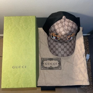 Gucci keps Tiger - Snygg keps från Gucci med ikoniskt GG-mönster och broderade tigrar framtill. Kepsen har en brun bas med svarta mesh-detaljer på sidorna och justerbar passform. Perfekt för att ge din outfit en lyxig touch.