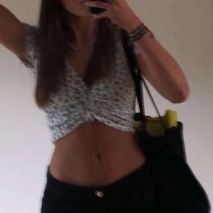 Vit blommig croppad topp - Säljer en söt vit croppad topp med blå blommor och knytning framtill. Toppen har korta ärmar och en v-ringad design. Perfekt för varma dagar!