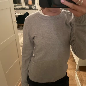 Grå stickad tröja - Säljer en stilren grå stickad tröja från vila  med rund halsringning. Tröjan har långa ärmar och ribbade muddar vid ärmslut och nederkant. Perfekt för kyligare dagar.