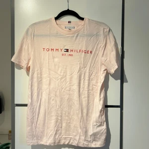 Ljusrosa t-shirt från Tommy Hilfiger - Säljer en stilren ljusrosa t-shirt från Tommy Hilfiger med röd logotyp. T-shirten är kortärmad och gjord i mjukt bomullsmaterial.