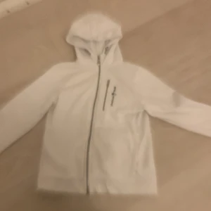 Vit kofta från Sail Racing (160) - Säljer en stilren vit jacka från Sail Racing med huva och dragkedja framtill. Perfekt för en sportig look. Jackan har långa ärmar och en enkel design som passar till mycket.