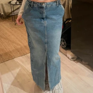 Jeanskjol från NA-KD - Snygg lång jeanskjol från NA-KD i klassisk blå denim. Kjolen har en stilren design med en slits framtill och knäppning i midjan. Perfekt för en trendig look!