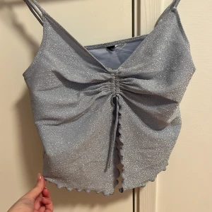 Glittrig silverfärgad linnetopp - Snygg glittrig linnetopp med smala axelband och v-ringning. Toppen har en rynkad detalj framtill och vågig nederkant som ger en unik look. Perfekt för att lysa upp vilken outfit som helst!