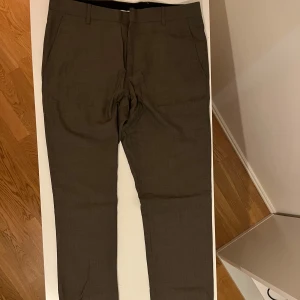 Bruna/grå kostymbyxor från Selected Homme - Snygga bruna kostymbyxor från Selected Homme. Byxorna har en klassisk design med knappar och dragkedja framtill. Perfekta för en stilren look.