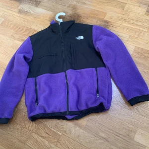 Lila fleecejacka från The North Face - Säljer en snygg lila fleecejacka från The North Face med svarta detaljer. Jackan har dragkedja framtill och praktiska fickor med dragkedjor. Perfekt för kyliga dagar och outdoor-äventyr. 🏞️