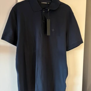 Helt ny! Mörkblå pikétröja från J. Lindeberg - Säljer en helt ny stilren mörkblå pikétröja från J. Lindeberg. Tröjan har en klassisk krage och knappar framtill. 