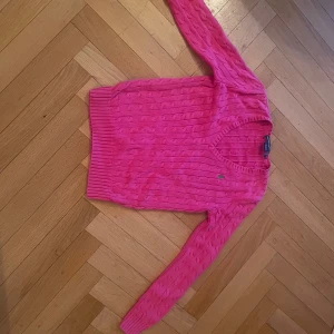 Rosa stickad tröja från Ralph Lauren - Säljer en snygg rosa stickad tröja från Ralph Lauren med kabelstickat mönster. Tröjan har en v-ringad hals och långa ärmar. Perfekt för att hålla sig varm och stilren. 💖