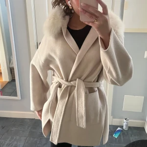 Beige kappa med päls luva - Säljer en beige kappa med en pälsdetalj runt huvan. Kappan har en knytning i midjan. Använd fåtal gånger.