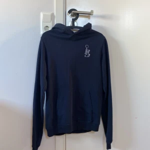 Mörkblå hoodie från Brand Store - Säljer en stilren mörkblå hoodie från Brand Store. Tröjan har en liten broderad figur på bröstet och en klassisk känguruficka framtill. Perfekt för en avslappnad look. (Passar mig som bär xs/s)