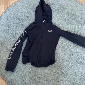 Svart hoodie från Emporio Armani - Snygg svart hoodie från Emporio Armani med dragkedja och huva. Den har märkets logga tryckt längs ena ärmen och en liten logga på bröstet. Perfekt för en stilren och bekväm look. Den är använd flera gånger men man ser inte det.Den är limited edition så svår att få tag på, denna är i storlek m-s. Kostar nu 400kr men kan gå ner mot 300kr i snabb affär+ frakt. 8/10. Skick