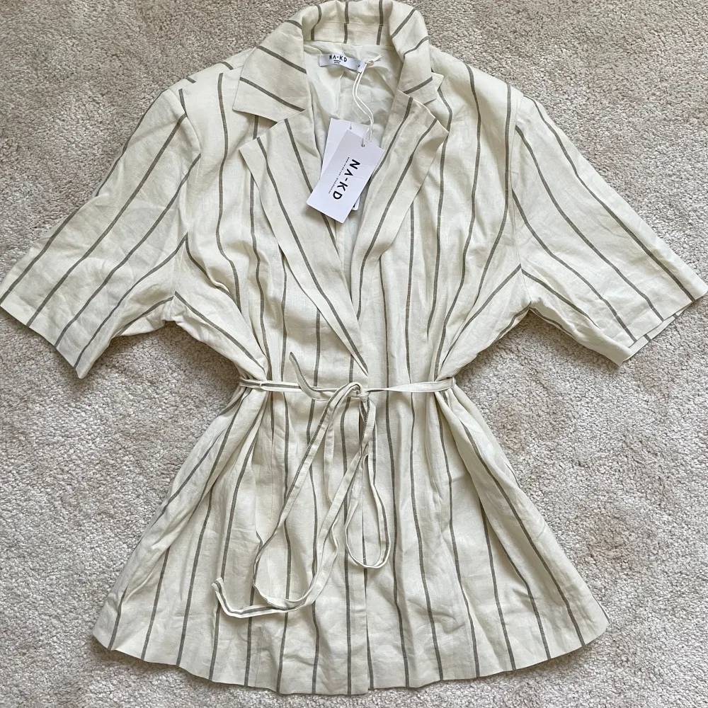 Snygg beige kavaj från NA-KD med tunna ränder och knytband i midjan. Perfekt för en stilren look. Kavajen har korta ärmar och en avslappnad passform.. Takit.