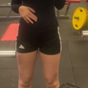 Svarta träningsshorts från Adidas - Snygga svarta träningsshorts från Adidas med vita ränder på sidorna. Perfekta för gymmet eller löprundan. De har en tight passform och är tillverkade i ett bekvämt syntetmaterial.