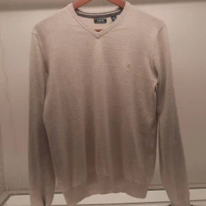 Beige tröja från IZOD - Säljer en stilren beige tröja från IZOD med v-ringning och ett diskret broderat emblem på bröstet. Tröjan har långa ärmar och är perfekt för en klassisk look. Passar bra till både jeans och chinos.