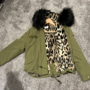 Olivgrön parkajacka med leopardfoder från ONLY - Snygg olivgrön parkajacka från ONLY med leopardmönstrat foder och svart fuskpäls på huvan. Jackan har dragkedja och knappar framtill samt justerbar midja för perfekt passform. Perfekt för att hålla stilen i kyligare väder!
