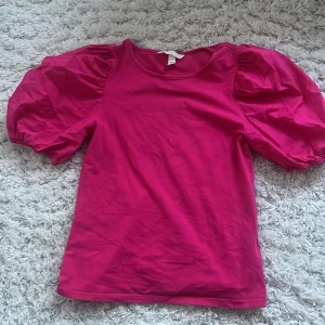 Rosa topp med puffärmar från H&M - Säljer en snygg rosa topp från H&M med puffiga kortärmar. Perfekt för att ge en färgklick till din outfit. Toppen har en rund halsringning och är gjord i ett mjukt material.