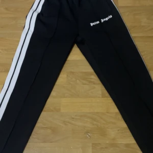 Svarta byxor från Palm Angels - Snygga svarta byxor från Palm Angels med vita ränder längs sidorna. Byxorna har en elastisk midja och är perfekta för en avslappnad stil. Logotypen är tryckt på framsidan.