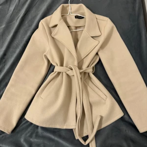 Beige kavaj med knytband - Elegant beige kavaj från Shein med knytband i midjan för en figursydd look. Kavajen har långa ärmar och klassisk krage, perfekt för en stilren outfit.