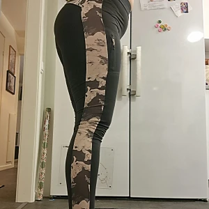 Svarta leggings med kamouflagedetaljer - Snygga svart-grå leggings med kamouflagemönstrade paneler längs sidorna. De har en praktisk ficka med dragkedja på sidan och är tillverkade i ett stretchigt material av polyester och elastan.