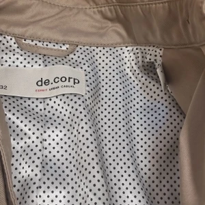 Beige trenchcoat från de.corp - Snygg beige trenchcoat från de.corp med klassisk design. Jackan har knappar framtill och ett bälte i midjan för en figursydd look. Insidan är fodrad med ett prickigt mönster i svart och vitt. Perfekt för vår och höst!