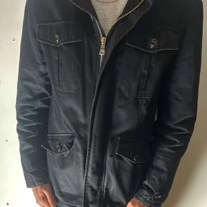 Svart jacka med dragkedja och knappar - Snygg svart jacka med dragkedja och knappar framtill. Jackan har flera fickor med lock och knappar, vilket ger en praktisk och stilren look. Perfekt för höst- och vårsäsongen.