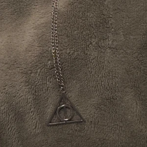 Silverfärgat halsband med deathly hallows berlock  - Snyggt silverfärgat halsband med deathly hallows berlock som har en cirkel och en linje inuti. Perfekt för att ge en stilren touch till din outfit.