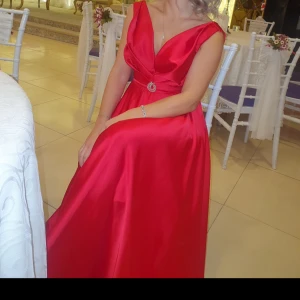 Röd långklänning med smyckesdetalj - Elegant röd långklänning i satin med djup v-ringning och smyckesdetalj i midjan. Klänningen har breda axelband och en vacker drapering framtill. Passar S och XS