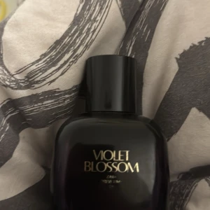 Violet Blossom parfym från Zara - Elegant parfymflaska i mörk lila med svart lock. Doften Violet Blossom från Zara ger en känsla av mystik och förförisk blomsterprakt. Perfekt för den som vill ha en sofistikerad och unik doftupplevelse. Är halva kvar