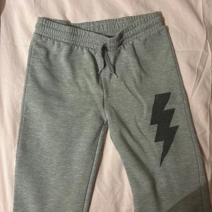 Grå mjukisbyxor med blixtdetalj - Säljer ett par grå mjukisbyxor med en cool svart blixtdetalj på benet. Byxorna har en elastisk midja med snörning för extra komfort. Perfekta för en avslappnad stil. Dem passar mig som är 1.62cm