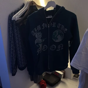 Mörkblå hoodie med dragkedja från Aim for the Moon - Snygg mörkblå hoodie med dragkedja och glittrigt tryck framtill. Perfekt för en avslappnad stil med en touch av bling. Huvan ger extra mysfaktor och ribbade muddar håller kylan ute.