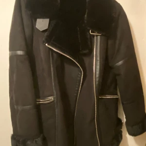 Svart jacka med pälsdetaljer - Snygg svart jacka med pälsdetaljer och dragkedja framtill. Jackan har en klassisk design med långa ärmar och en mjuk krage. Perfekt för kyligare dagar.