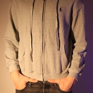 Grå ziphoodie från Ralph Lauren - Säljer en stilren grå ziphoodie från Ralph Lauren med dragkedja och huva. Perfekt för en avslappnad look. Den har en liten broderad logga på bröstet och ribbade muddar.