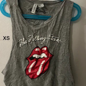 Grå Rolling Stones linne och shorts - Säljer ett grått linne och matchande shorts med Rolling Stones ikoniska tunga-tryck. Linnet är ärmlöst och shorts har ett elastiskt band i midjan. Perfekt för en avslappnad stil.
