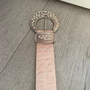 Rosa skärp med glittrigt spänne - Snyggt rosa skärp i äkta läder från Saddler. Skärpet är 80 cm långt.