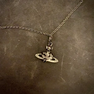 Vivienne Westwood Halsband - Stilrent silverfärgat halsband från Vivienne Westwood med ett unikt hängsmycke. Kedjan är tunn och elegant, perfekt som julklapp till både män och kvinnor.