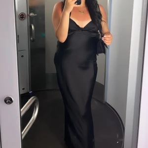 Svart långklänning - Elegant svart långklänning med en v-ringad design och smala axelband. Klänningen har en figurnära passform som framhäver silhuetten. Perfekt för en stilren och sofistikerad look.